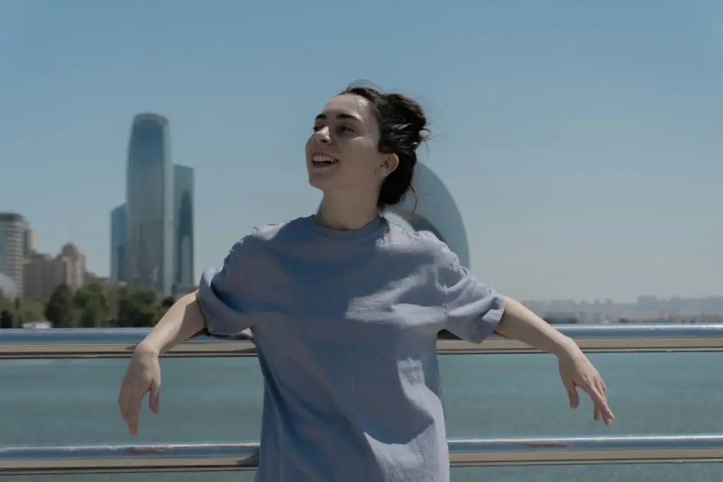 A woman stands outdoors by the water with a city skyline behind her, arms relaxed and open, taking in a moment of lightness, calm, and emotional freedom. It's not that bad - therapy in Plano Texas